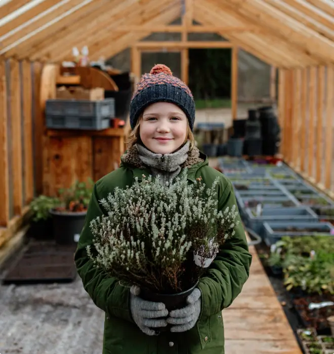 Un enfant souriant tenant une plante en pot dans une serre en bois, illustrant l'approche éducative et la sensibilisation à la nature à la Vallée de la Millière.